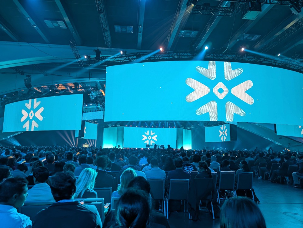 Große Konferenz mit vielen Teilnehmern, beleuchtet von großen Bildschirmen mit einem Schneeflocken-Logo.