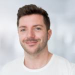 Ein lächelnder Mann mit kurzen, braunen Haaren trägt ein weißes T-Shirt vor unscharfem Hintergrund.