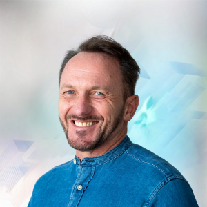 Mann mit kurzem Bart und lächelndem Gesicht, trägt ein blaues Hemd, vor einem sanften, abstrakten Hintergrund.