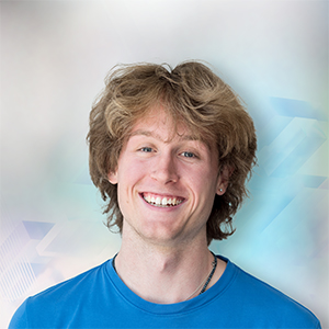 Junger Mann mit lockigem, blondem Haar und lächelndem Gesicht, trägt ein blaues T-Shirt vor einem verschwommenen Hintergrund.
