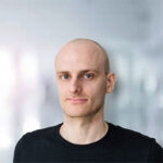 Ein Mann mit glattem Kopf und schwarzem T-Shirt vor einem unscharfen Hintergrund.