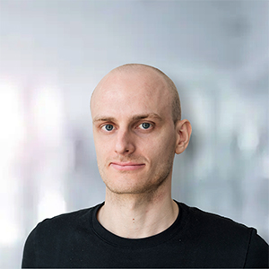Ein Mann mit glattem Kopf und schwarzem T-Shirt vor einem unscharfen Hintergrund.