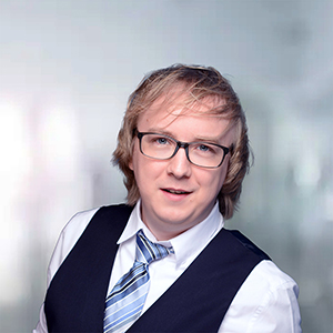 Ein junger Mann mit Brille, blonden Haaren und Anzugweste, lächelt vor einem unscharfen, hellen Hintergrund.