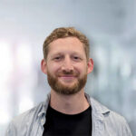 Ein Mann mit blonden Haaren und Bart lächelt vor einem unscharfen, hellen Hintergrund.