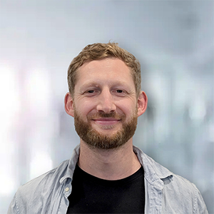 Ein Mann mit blonden Haaren und Bart lächelt vor einem unscharfen, hellen Hintergrund.