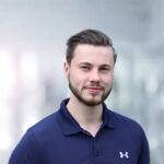 Ein junger Mann mit Bart und kurzen Haaren, trägt ein blaues Poloshirt, lächelt vor einem unscharfen Hintergrund.