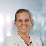 Frau mit lächelndem Gesicht, in blasser Bluse und Blazer, vor verschwommenem Hintergrund.