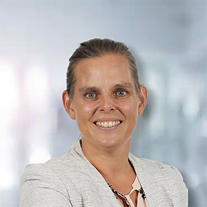 Frau mit lächelndem Gesicht, in blasser Bluse und Blazer, vor verschwommenem Hintergrund.