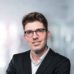 Ein junger Mann mit Brille in einem schwarzen Anzug vor einem unscharfen Hintergrund.