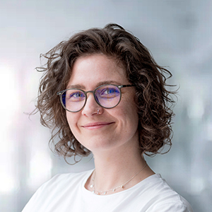 Eine lächelnde Frau mit lockigem Haar und Brille, vor einem unscharfen Hintergrund.