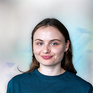Eine junge Frau mit langen braunen Haaren trägt ein blaues T-Shirt und lächelt vor einem unscharfen, hellen Hintergrund.