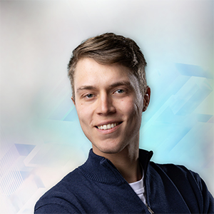 Junger Mann mit kurzem Haar, lächelnd vor einem verschwommenen, hellen Hintergrund. Trägt einen blauen Pullover.
