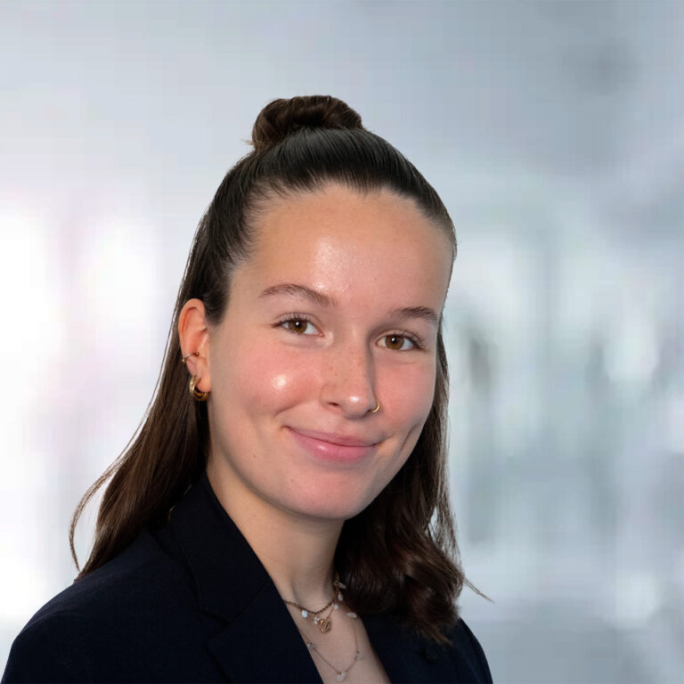 Porträt einer jungen Frau mit schulterlangen, braunen Haaren, die in einem eleganten Blazer lächelt, vor unscharfem Hintergrund.
