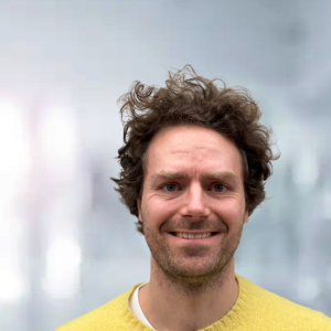 L&auml;chelnder Mann mit lockigem Haar in einem gelben Pullover, vor unscharfem Hintergrund.