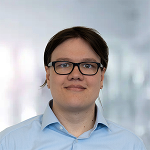 Person mit Brille und kurzem, dunklem Haar in einem hellblauen Hemd vor unscharfem Hintergrund.