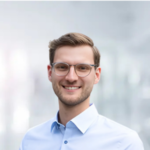 Ein junger Mann mit Brille lächelt vor unscharfem Hintergrund, trägt ein hellblaues Hemd.