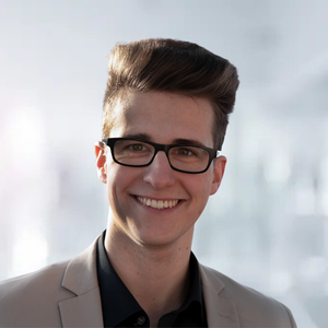 Lächelnder junger Mann mit Brille und modernem Haarschnitt, trägt einen hellen Anzug vor unscharfem Hintergrund.
