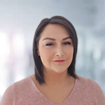 Eine Frau mit schulterlangen, braunen Haaren und einem neutralen Ausdruck trägt ein rosa Oberteil.