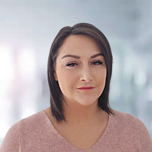 Eine Frau mit schulterlangen, braunen Haaren und einem neutralen Ausdruck trägt ein rosa Oberteil.