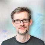Ein Mann mit grauen Haaren und Brille lächelt vor einem unscharfen, abstrakten Hintergrund.