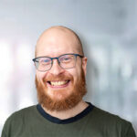 Lächelnder Mann mit Brille und Bart, vor einem unscharfen hellen Hintergrund.