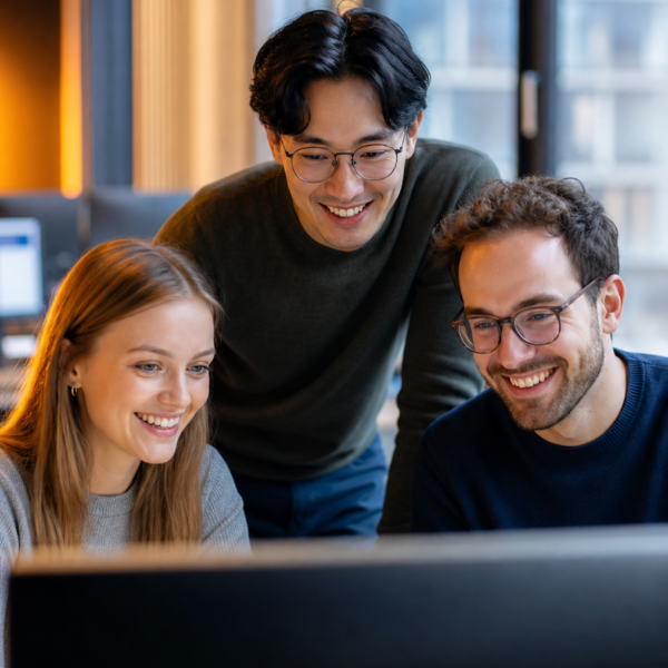 Drei Personen l&auml;cheln und schauen gemeinsam auf einen Computerbildschirm in einem modernen B&uuml;ro.