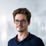 Junger Mann mit lockigem Haar und Brille, lächelnd vor verschwommenem, hellem Hintergrund.