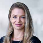 Porträt einer Frau mit langen blonden Haaren und blauen Augen, lächelnd vor einem hellen, unscharfen Hintergrund.