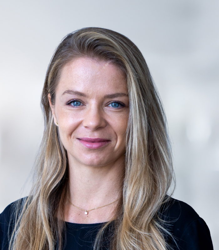 Porträt einer Frau mit langen blonden Haaren und blauen Augen, lächelnd vor einem hellen, unscharfen Hintergrund.