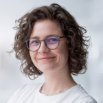 Porträt einer lächelnden Frau mit lockigem Haar, Brille und einem weißen T-Shirt, vor einem unscharfen Hintergrund.