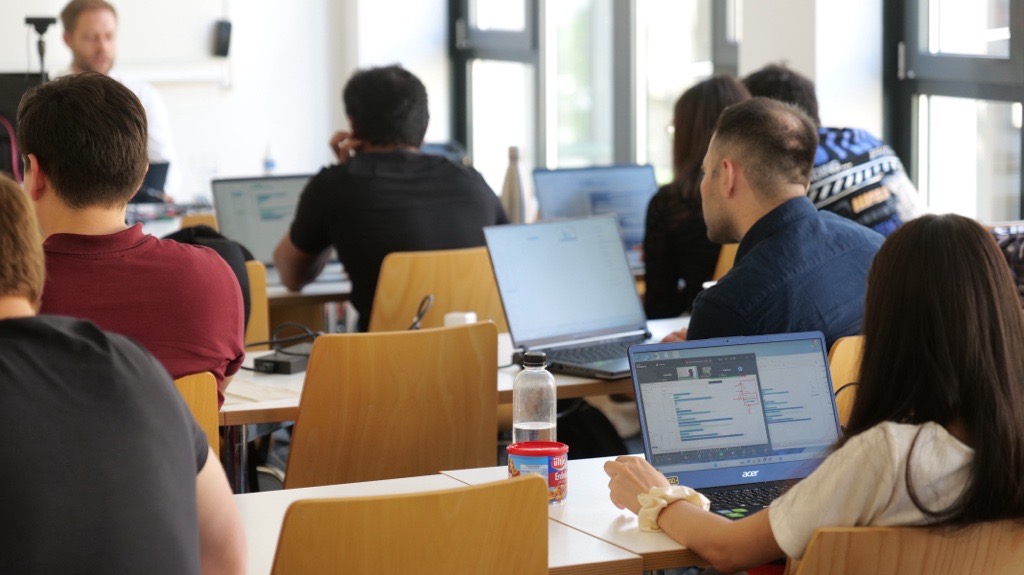 Studierende in einem Klassenzimmer, arbeiten an Laptops, während ein Dozent vorne präsentiert.
