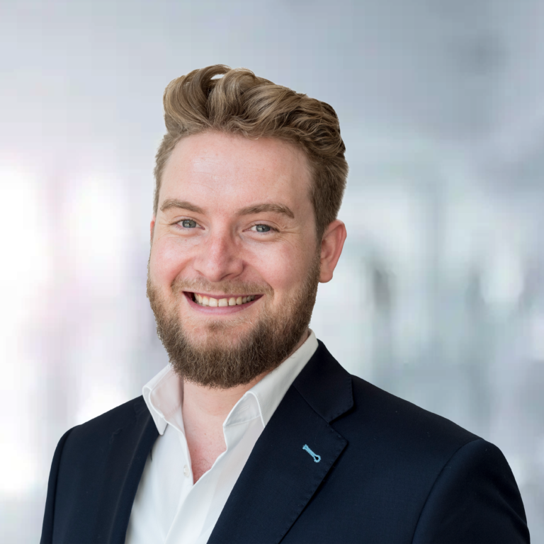 Ein lächelnder Mann mit Bart in einem Anzug vor einem unscharfen, hellen Hintergrund.